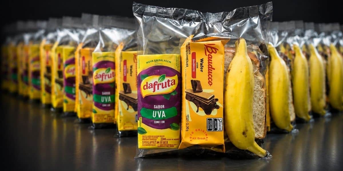 Fileira de Kit Lanches embalados individualmente em Flow Pack, com suco, wafer, pão e banana, lacrados e padronizados